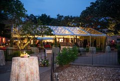Cocktail tables with floral and sage linens placed in the gardens outside of an outdoor lighted dance tent