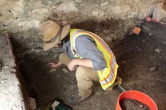 Archaeologist wearing hat and vest cleaning wall inside excavation unit