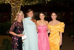 Four ladies in gowns at an evening outdoor event smiling by a tree wrapped in string lights