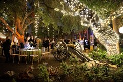 String lights hanging from trees and oak wrapped in string lights at an outdoor event in Alamo Gardens with tables and a demonstration cannon